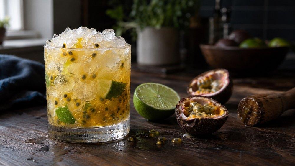Caipirinha s marakujou v nízkom pohári s drveným ľadom, dužinou marakuje so semienkami a kúskami limetky, vedľa dve polovice marakuje, polovica limetky a drevený muddler.