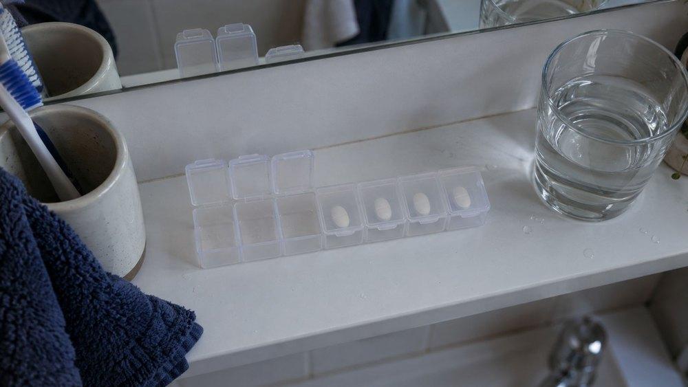 A weekly pill organiser on a bathroom shelf, with three compartments already open and empty and one tablet in each of the four remaining compartments, next to a glass of water and a toothbrush cup.