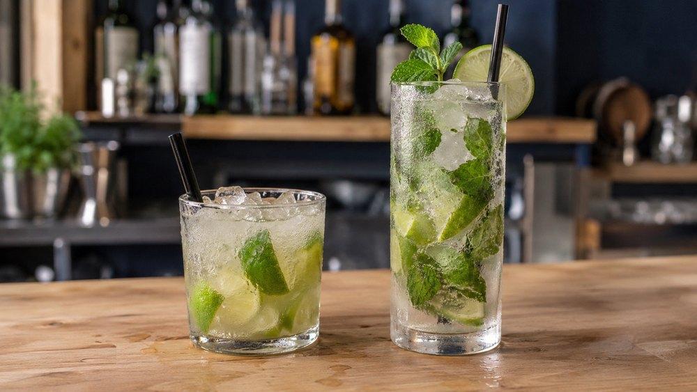 Dva drinky vedľa seba na barovom pulte: vľavo Caipirinha v nízkom širokom pohári s drveným ľadom a limetkou, vpravo Mojito vo vysokom pohári s ľadom, mätou a limetkou.
