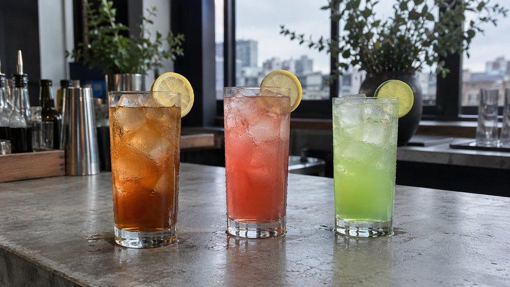 Tři vysoké sklenice s ledem vedle sebe na betonovém barovém pultu: jantarově hnědý Long Island s citronem, růžový Long Beach s citronem a světle zelený Tokyo Iced Tea s limetkou.