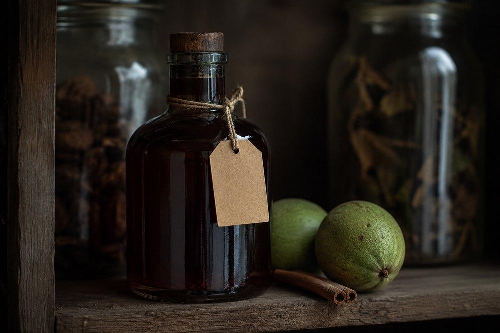 Dark glass bottle of finished walnut liqueur with a paper label on a wooden pantry shelf, accompanied by green walnuts and cinnamon.