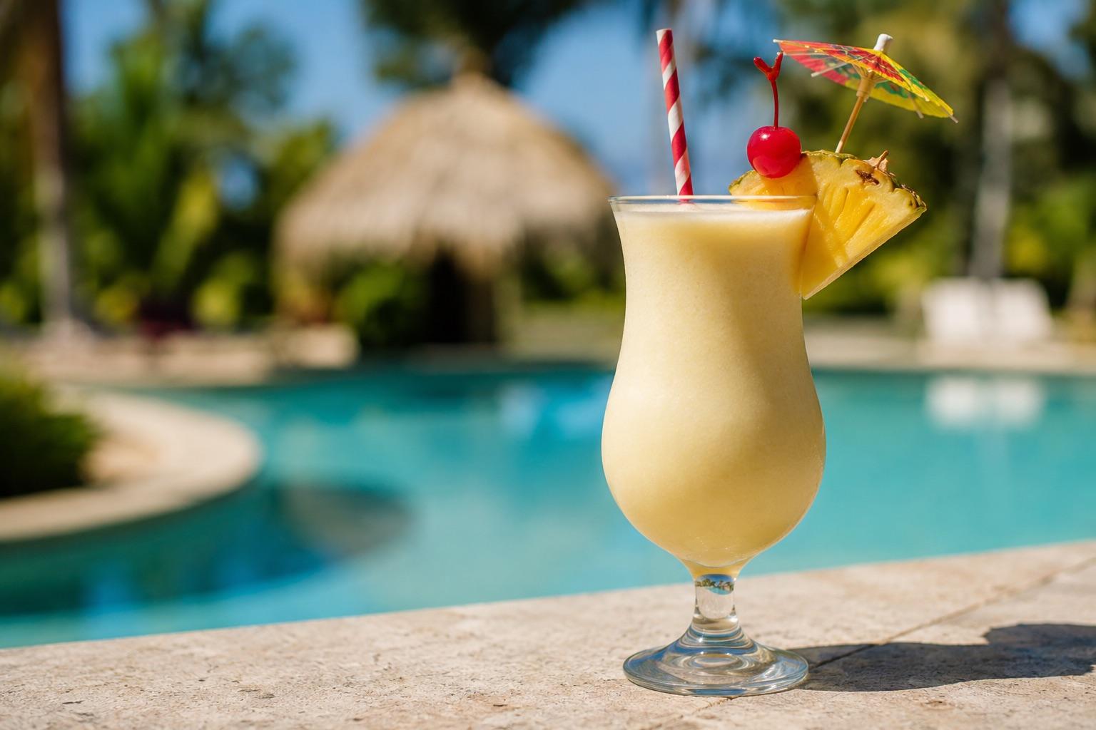 Piña Colada served in a hurricane glass with a paper umbrella, straw, pineapple and cherry beside a swimming pool.