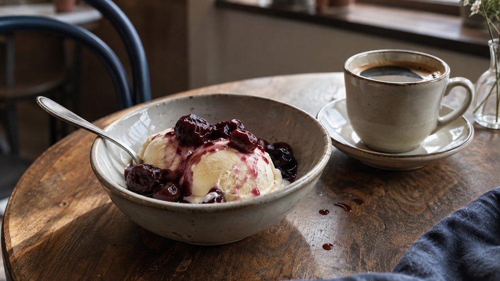 A bowl of vanilla ice cream topped with sour cherries from homemade Griotka and dark cherry syrup, beside a cup of black coffee on a wooden café table.